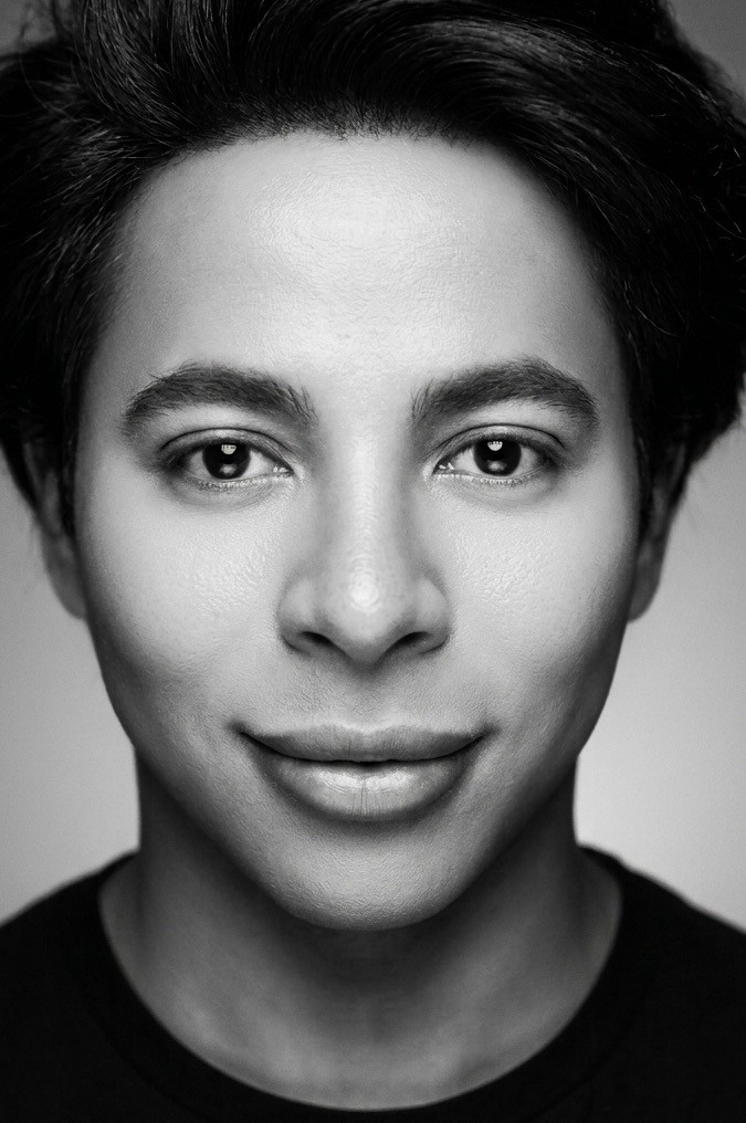 Close-up black-and-white portrait of a person with short dark hair and a neutral expression, looking at the camera.